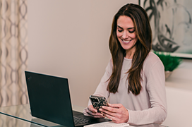 Smiling woman sitting in front of a laptop looking at her mobile device.