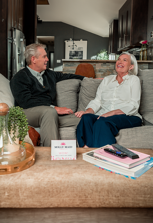 Smiling senior couple sitting on their couch together behind a Molly Maid promotional card.