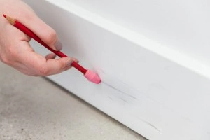 A hand erasing scuff marks on a white baseboard with a pencil eraser