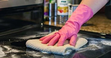 Person wearing a pink rubber glove cleaning a black stovetop with a cloth.