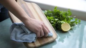 A person wiping a wood cutting board with a cloth, next to a sliced lime and bunch of parsley