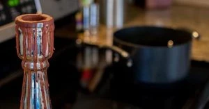 A candlestick covered in wax drippings in the foreground, with a pot on a stovetop in the background