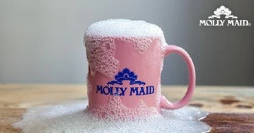 A sudsy pink coffee mug with the Molly Maid logo on a wood table.