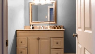 Door opening into bathroom with pale blue walls and mirror in gold frame hanging over sink cabinet.