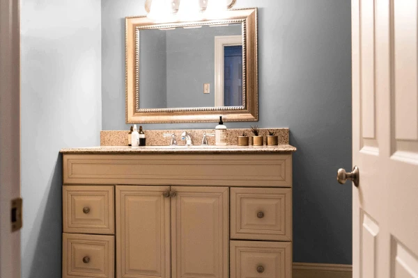 Door opening into bathroom with pale blue walls and mirror in gold frame hanging over sink cabinet.