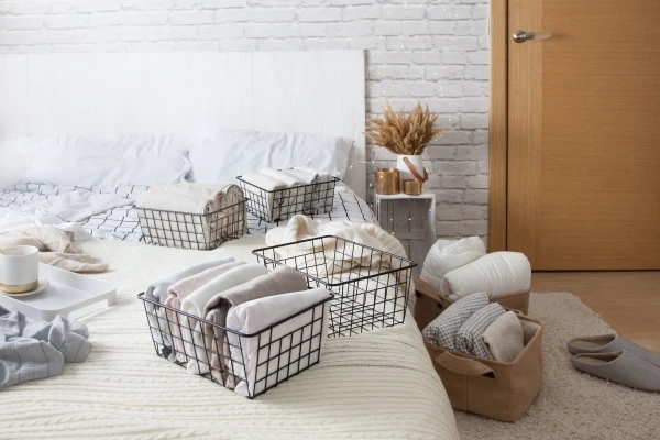 Mesh metal containers full of clothes and linens on the bed and floor in a bedroom. Cup of coffee on a serving tray on the bed.