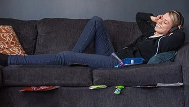 Woman smiling and lying on a couch, with headphones on and food wrappers stuck under the cushions