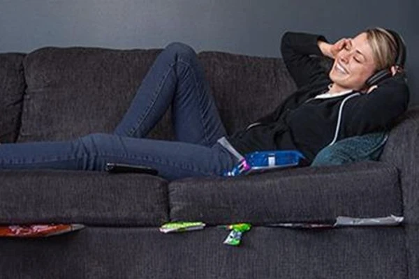 Woman smiling and lying on a couch, with headphones on and food wrappers stuck under the cushions