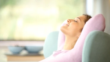 Woman with arms folded behind her head and eyes closed relaxing on her couch at home.