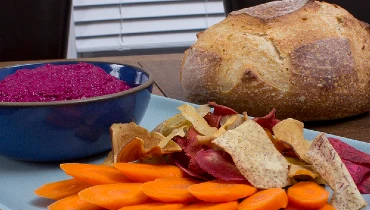 A gray serving tray with beet hummus, carrots, and veggie chips in front of a loaf of bread