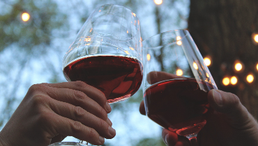 Two wine glasses containing red wine being clinked together in an outdoor setting, photo by rikkia hughes on Unsplash