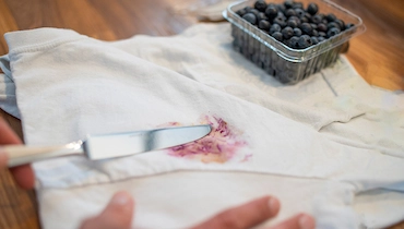 Berry stain on white cloth napkin with silver knife and pint of blueberries spread out on table.