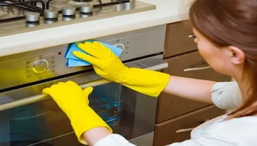 Woman wearing yellow rubber gloves cleaning oven exterior with blue cloth.