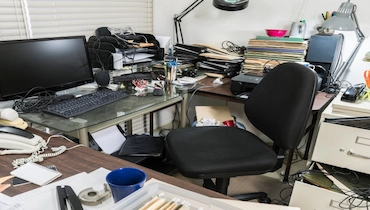 Desk of a home office covered in files and surrounded by clutter.