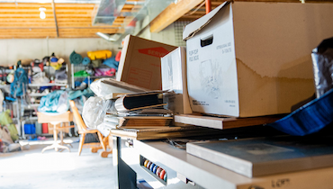 Carboard boxes and miscellaneous items piled on table and shelves inside cluttered garage.