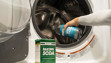 Person pouring white vinegar into washer with box of baking soda in foreground.