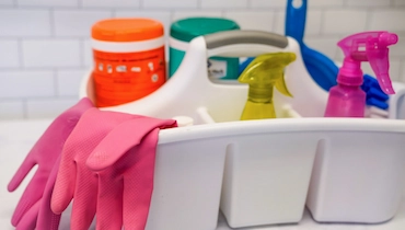 Pink rubber gloves and assorted colorful cleaning products in white plastic cleaning caddy.