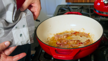 Close-up of gray checkered shirt with grease stains held up beside red pan with bacon grease on stovetop.