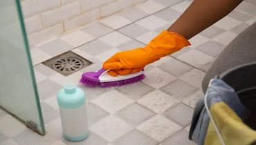 Person wearing orange rubber glove scrubbing shower floor with brush.