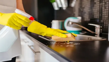 Person wearing yellow rubber gloves cleaning black countertop with spray and yellow sponge.