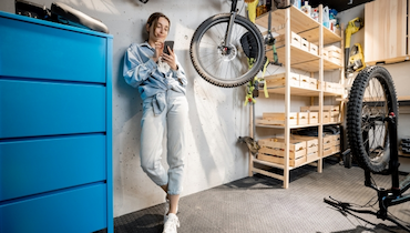 Young woman on her phone standing in a garage with bicycle hanging from ceiling.