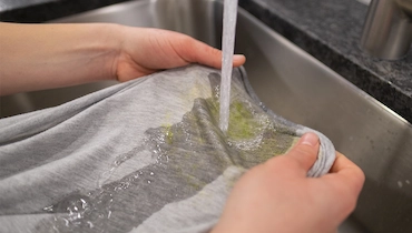 Person holding shirt with grass stains under running kitchen faucet.