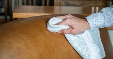 A hand wiping a tan leather couch with a cloth