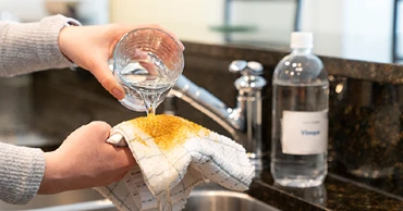 Water being poured from a glass onto a white cloth stained with turmeric, with a bottle of vinegar in the background.